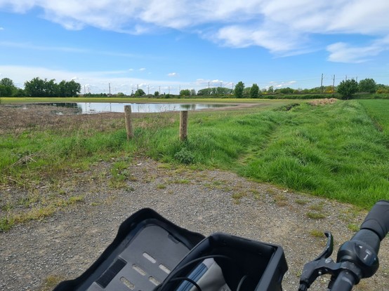 Landschaft mit Wiese, Acker, im Hintergrund Gleise, unten eine Fahrradtasche, auf dem Acker eine Pfütze